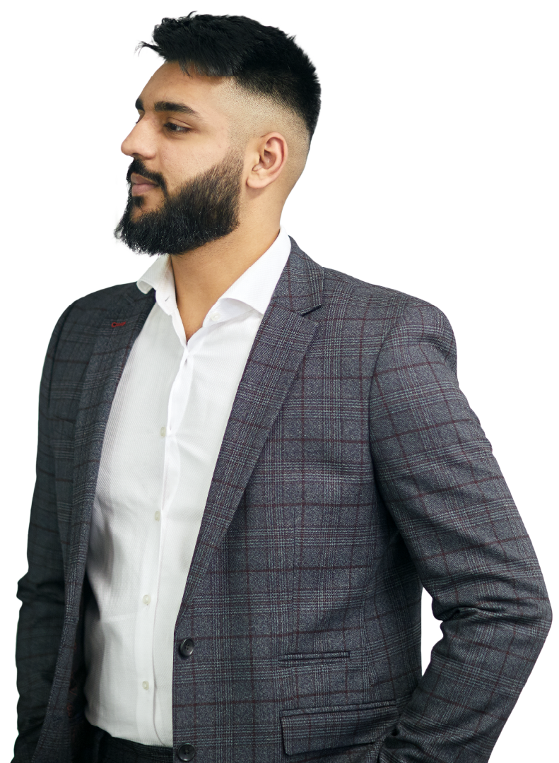 Bearded man wearing a white shirt and grey plaid blazer looks left with hands in pockets, standing against a plain light background; composed, confident posture.